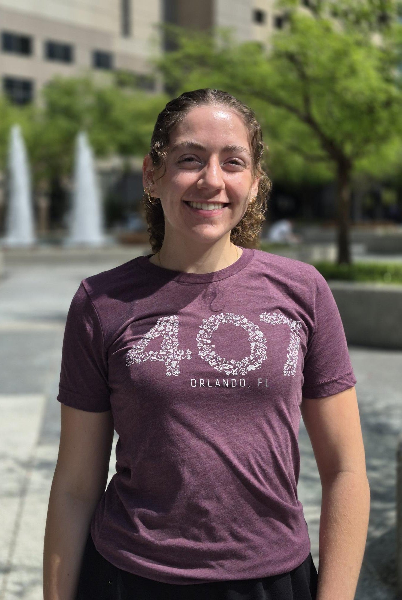 Person wearing a purple t-shirt with '407 Orlando, FL' text in an outdoor setting with fountains and trees.