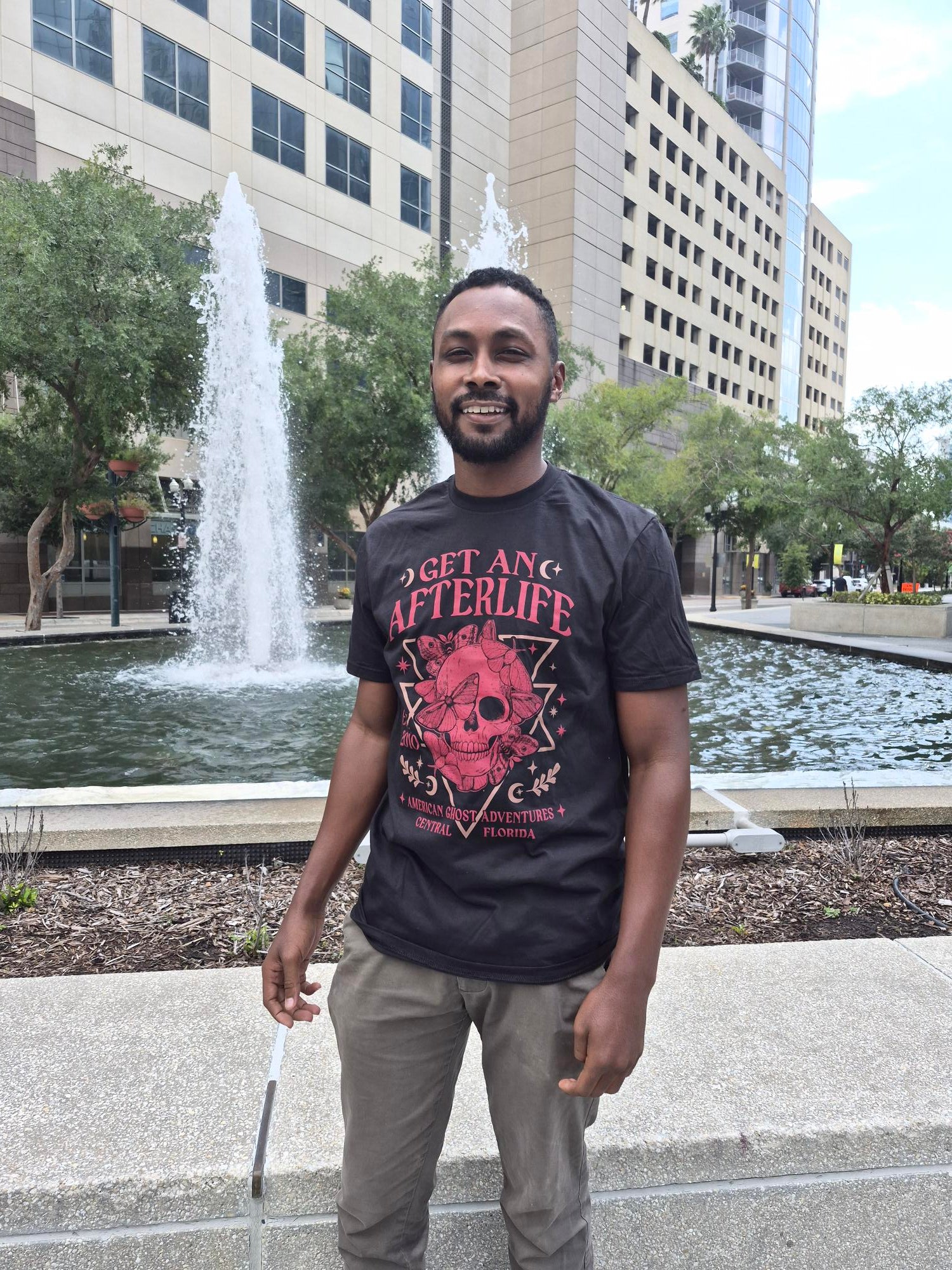 A man wearing a Black American Ghost Adventures Tee shirt in front of a fountain.