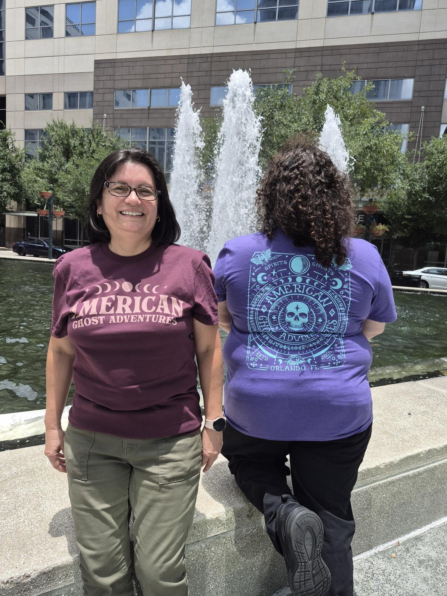 Two people wearing t-shirts with visible text standing in front of a fountain.