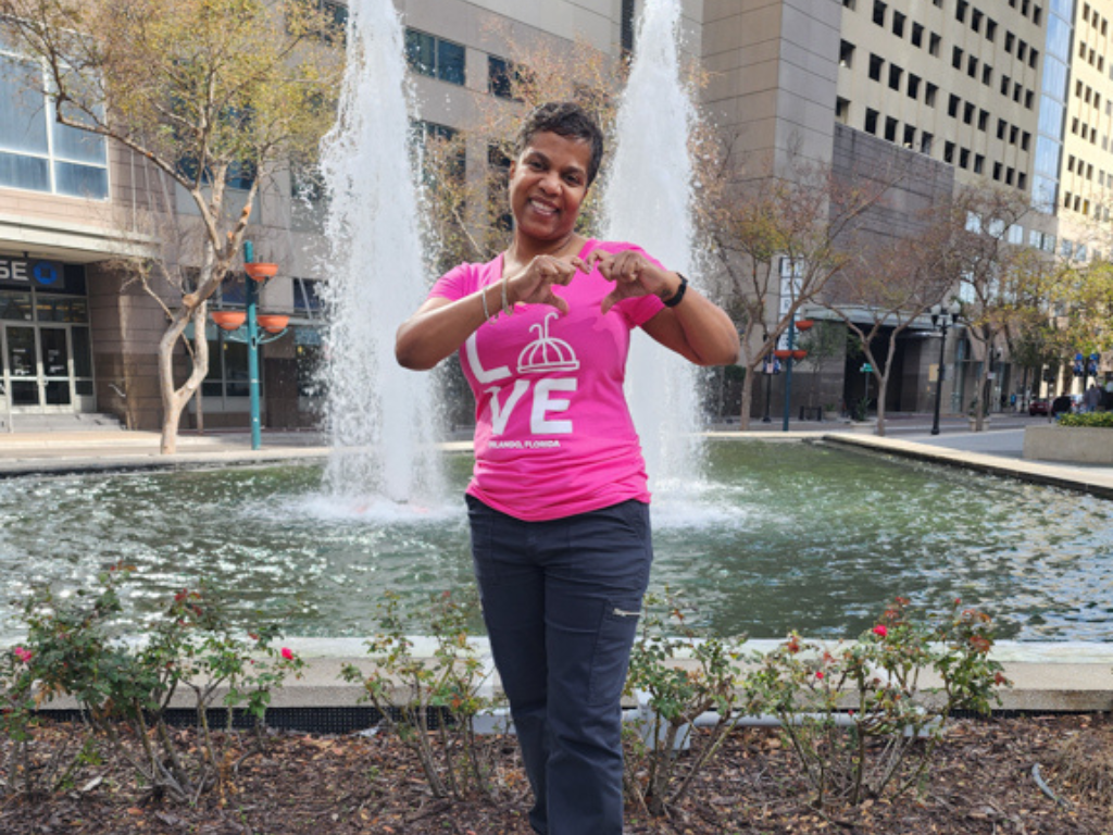 Person wearing a pink shirt with a logo and 'LOVE' text, standing in front of a fountain in an urban setting.