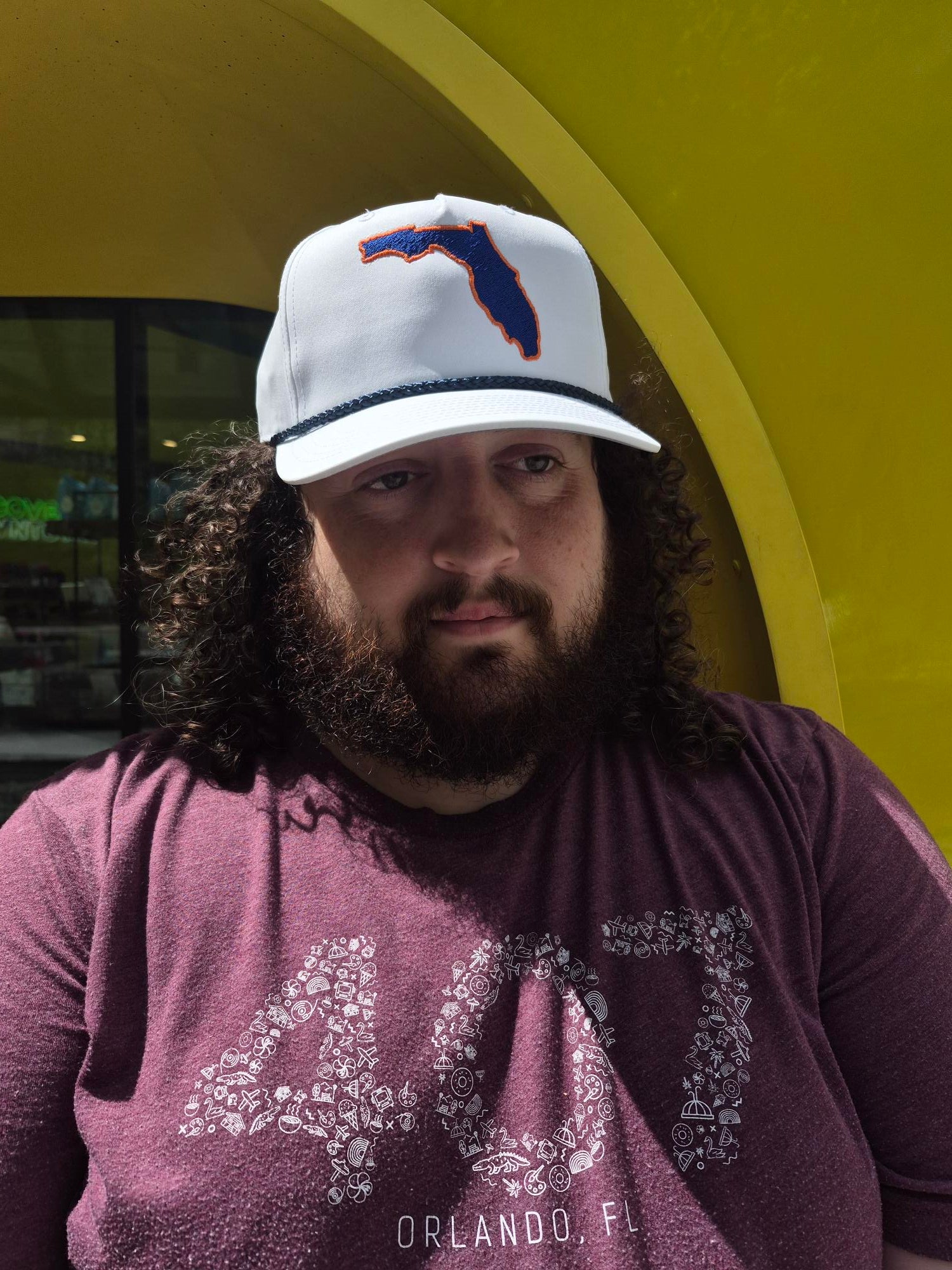 A man with curly hair and a beard wearing a white baseball cap with a blue and orange Florida state outline logo, paired with a mauve-purple Orlando shirt, photographed in a low-angle perspective against a yellow background.