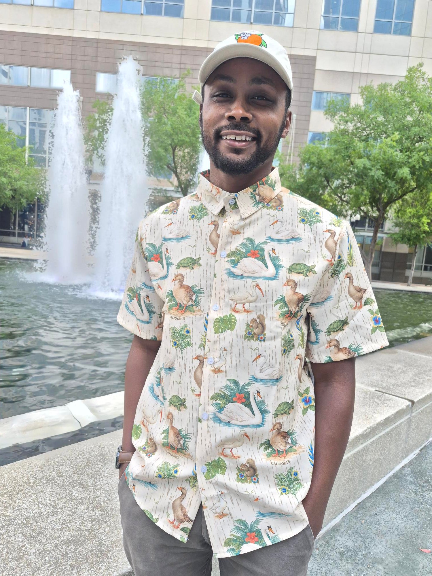 Person wearing a patterned shirt standing outdoors with a fountain in the background