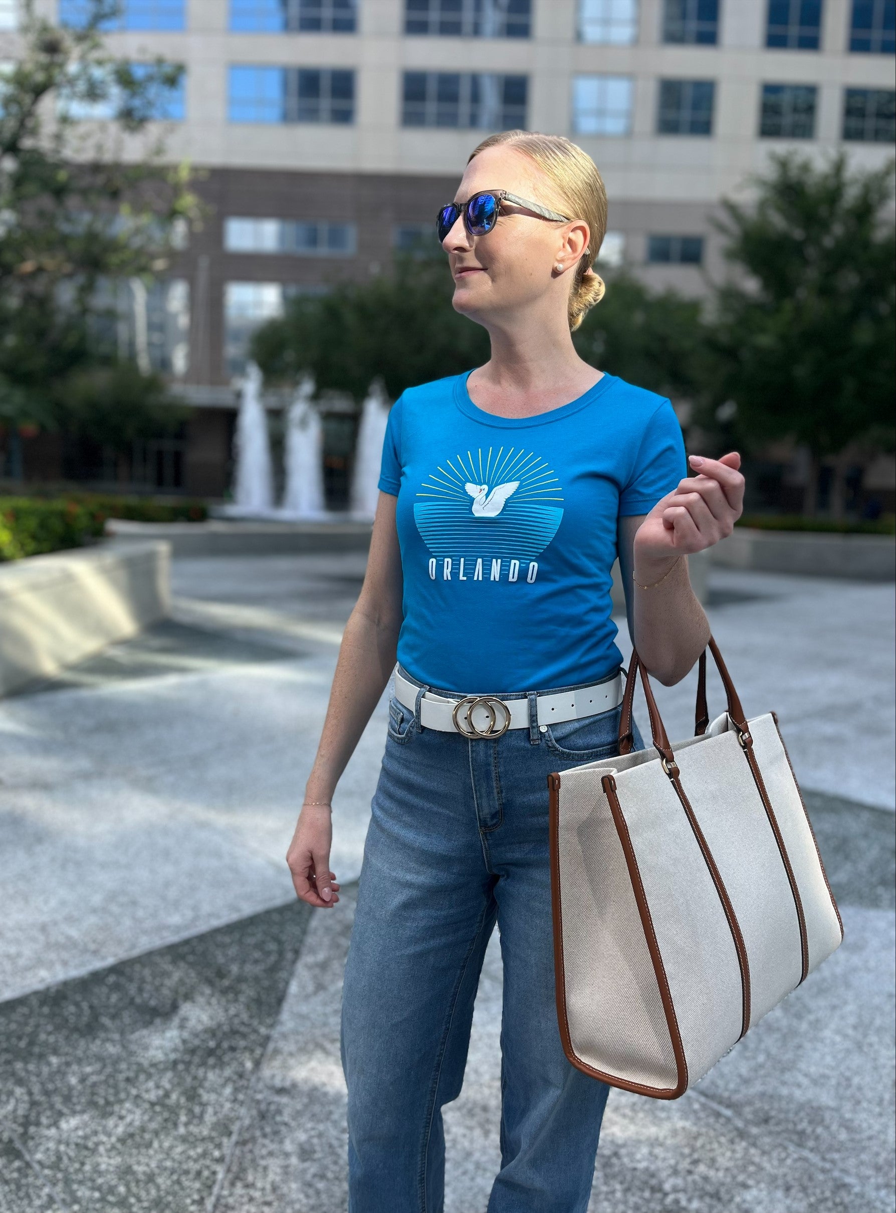 Woman wearing a blue t-shirt with a logo and text, holding a beige tote bag, standing outdoors.