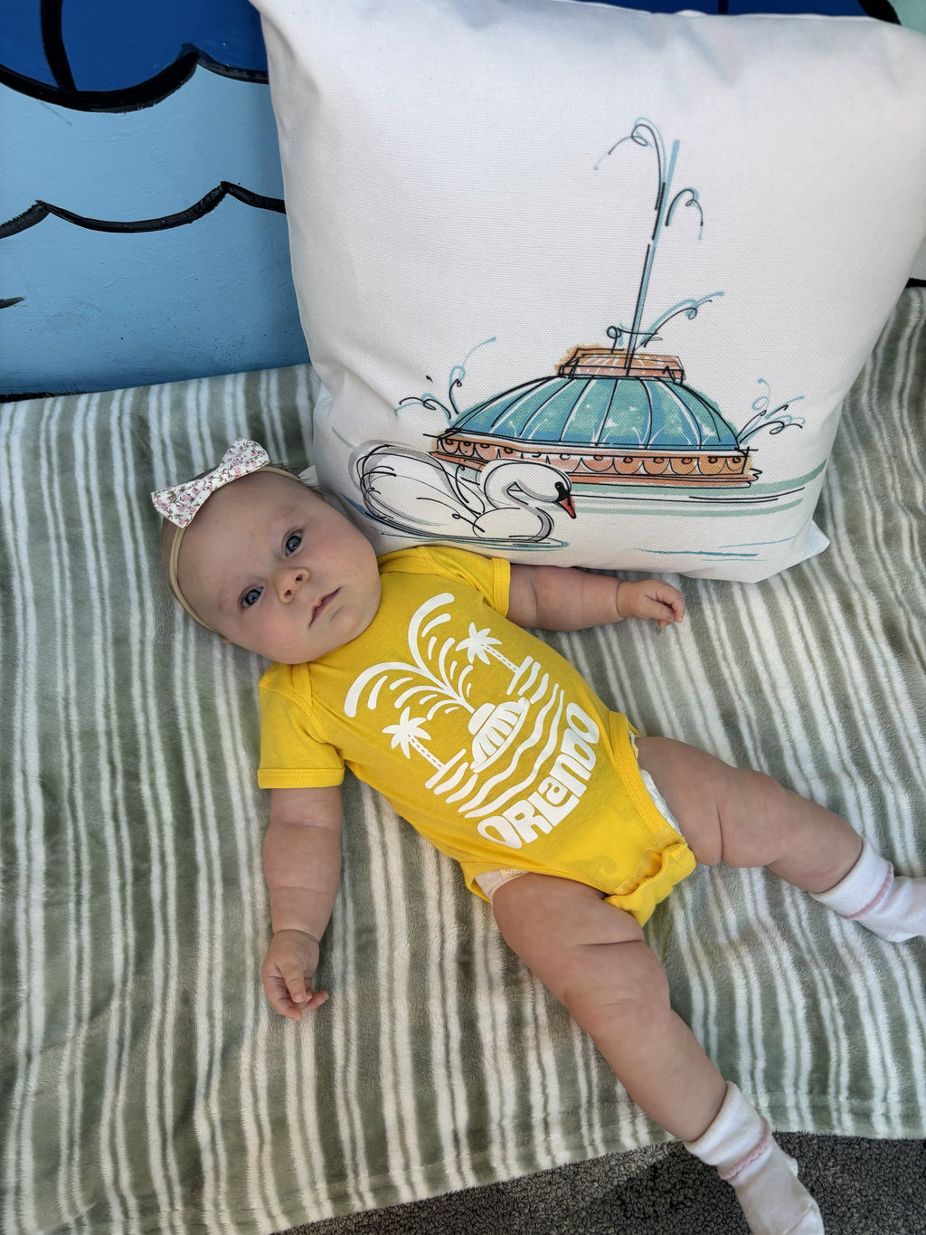 Baby in a yellow onesie lying on a striped blanket with a decorative pillow in the background.